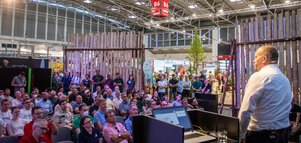 Eine große Anzahl von Zuhörern blickt hinauf auf eine Bühne in der Halle B6, auf der ein Mann neben einem Rednerpult eine Präsentation hält.