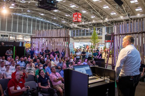  Eine große Anzahl von Zuhörern blickt hinauf auf eine Bühne in der Halle B6, auf der ein Mann neben einem Rednerpult eine Präsentation hält.