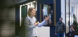 Eine Frau mit blauer Bluse und Brille spricht gestikulierend auf einer Bühne und hält dabei eine Fernbedienung in der anderen Hand.