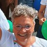 Lächelnde Person mit kurzen, lockigen grauen Haaren, Brille und einer Regenbogennadel, die im Freien einen grünen Ballon hält.