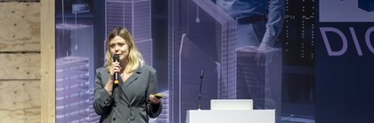 Junge Frau steht auf einer Bühne neben einem weißen Sprecherpult, auf dem ein digitalBAU Logo aufgebracht ist.