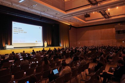 Ein großer Konferenzsaal mit sitzenden Teilnehmern vor einer Leinwand, auf der eine Präsentation läuft.