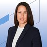 Frau mit dunklen Haaren im Business-Outfit vor blau-weißem, geometrischem Hintergrund; sie blickt freundlich in die Kamera.