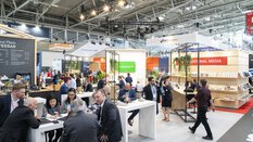 Besucher der transport logistic sitzen einer Kaffeebar in einer Messehalle und unterhalten sich, während im Hintergrund weitere Leute zwischen den Ständen entlang gehen.