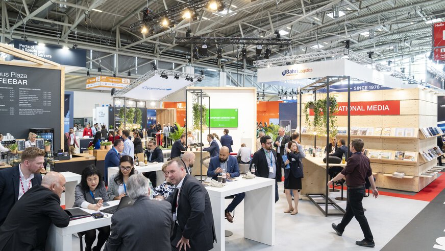 Besucher der transport logistic sitzen einer Kaffeebar in einer Messehalle und unterhalten sich, während im Hintergrund weitere Leute zwischen den Ständen entlang gehen.