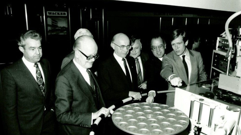 Black and white photo from an earlier productronica. Several men in suits and some with glasses are standing around an apparatus and looking at it.