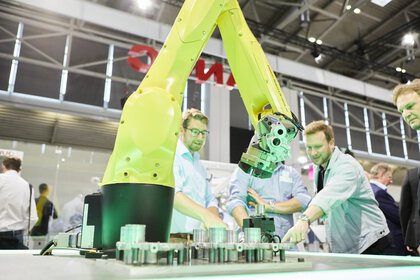 A yellow robot arm at a trade fair stand, with some interested trade fair visitors in the background.