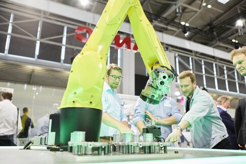 Ein gelber Roboterarm an einem Messestand, im Hintergrund sind einige interessierte Messebesucher zu sehen.