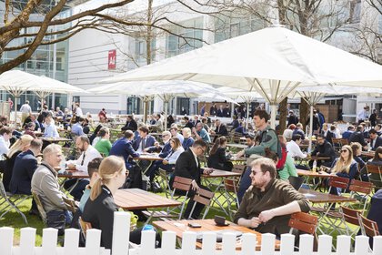 People sit at outdoor tables and enjoy a sunny day on a green area between the exhibition halls.