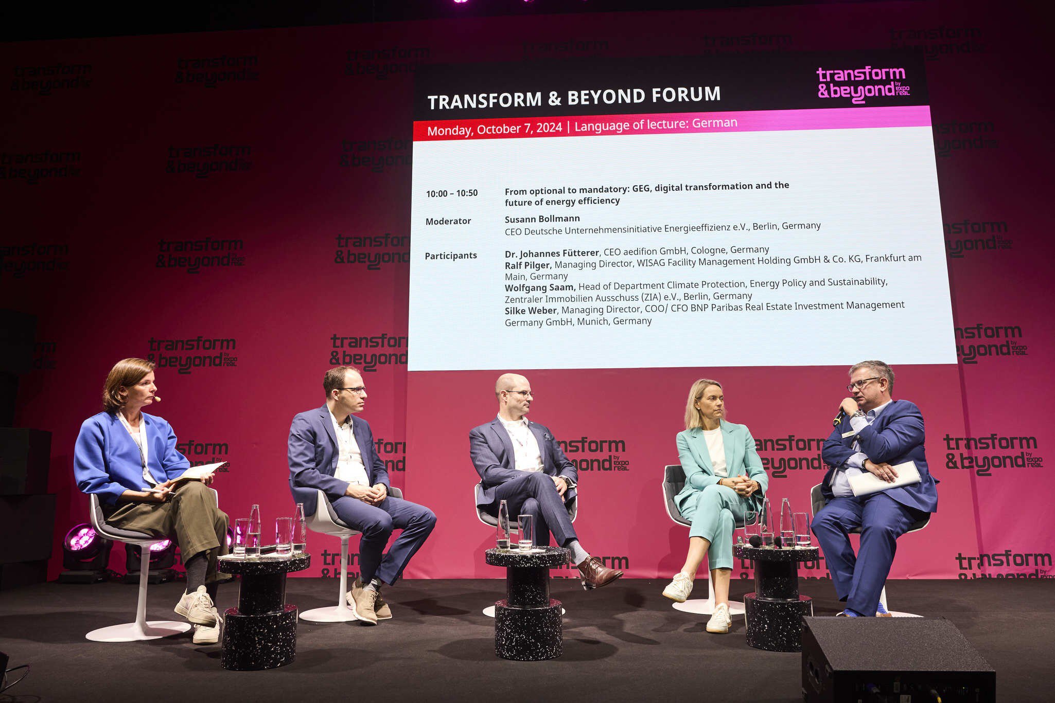Five people sit on the stage during a panel discussion in the Transform & Beyond Forum. Details of the discussion are displayed on the screen behind them.