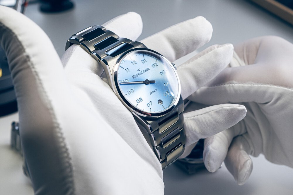 Zwei Hände in weißen Handschuhen halten eine silberne „Meistersinger“-Uhr mit einem blau schimmernden Ziffernblatt und mit einer eingelassenen Datumsanzeige.
