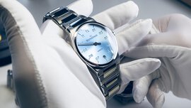Zwei Hände in weißen Handschuhen halten eine silberne „Meistersinger“-Uhr mit einem blau schimmernden Ziffernblatt und mit einer eingelassenen Datumsanzeige.