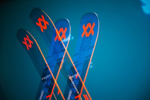 Drei gekreuzte Ski der Marke Völkl, zeigen leuchtend orange und blaue Designs vor einem auffälligen blaugrünen Hintergrund.