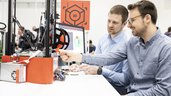 Zwei Männer sitzen an einem Tisch vor einem 3D-Drucker und einem Computer Bildschirm mit angezeigten Grafiken.