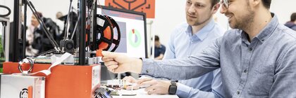 Zwei Männer sitzen an einem Tisch vor einem 3D-Drucker und einem Computer Bildschirm mit angezeigten Grafiken.