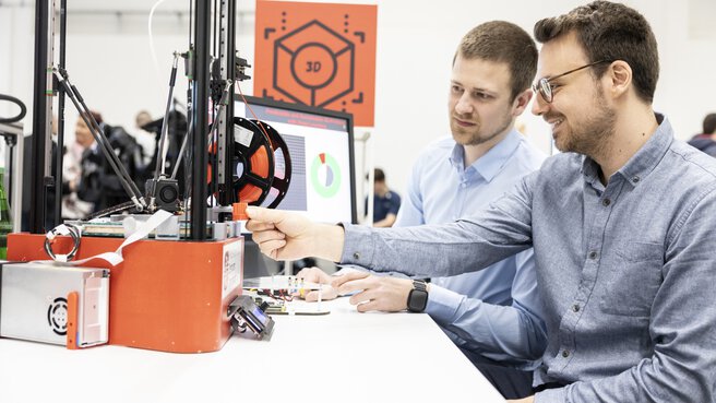 Zwei Männer sitzen an einem Tisch vor einem 3D-Drucker und einem Computer Bildschirm mit angezeigten Grafiken.