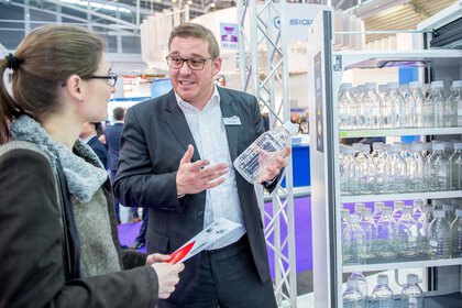 Auf der analytica zeigt ein Aussteller einer Besucherin eine Labor-Glasflasche und steht dabei neben einem Schrank gefüllt mit mehreren Labor-Glasflaschen.
