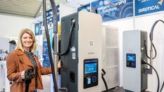 Eine blonde Frau steht links neben einer großen Ladesäule von connectika für Elektrofahrzeuge und lächelt in die Kamera.