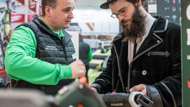 A trade fair visitor in a black suit, beard and hat examines tools at a trade fair stand while an exhibitor talks to him.