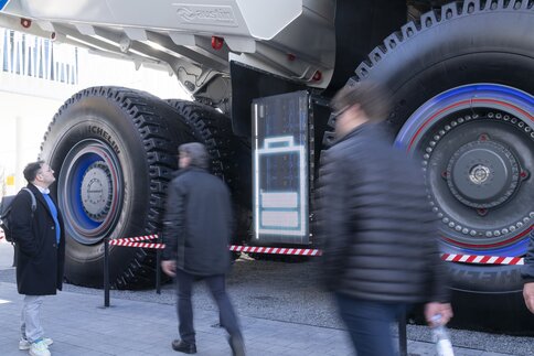 Several people walk past a large battery display between two giant wheels of a vehicle.