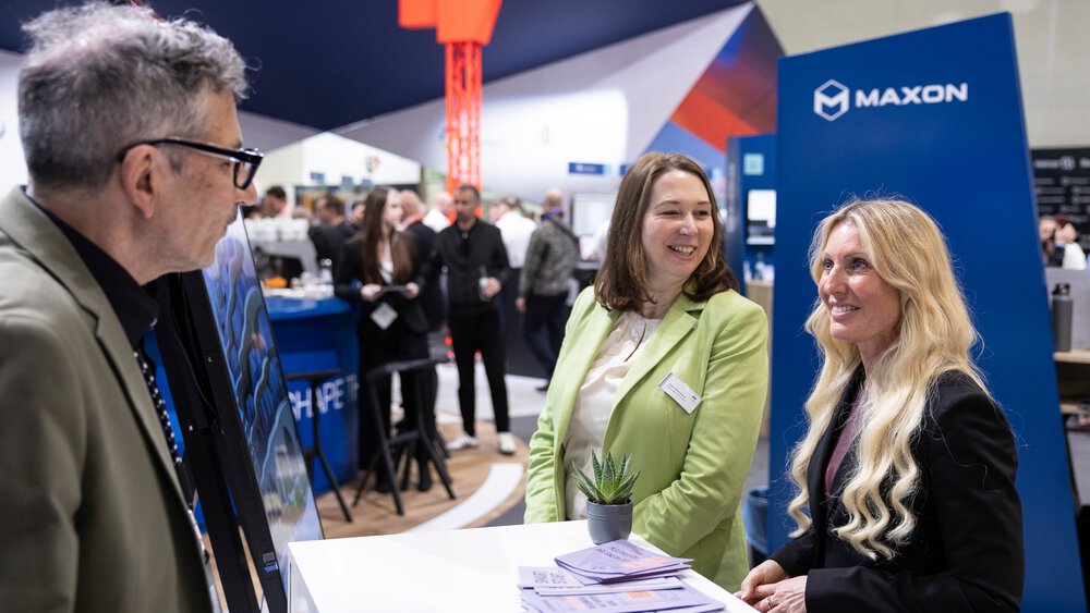 Zwei Frauen und ein Mann stehen an einem Tresen auf der digitalBAU. Im Hintergrund sind weitere Personen und ein orangener Kran zu sehen.