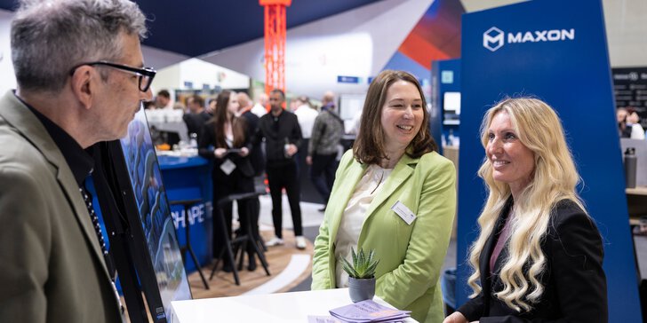 Zwei Frauen und ein Mann stehen an einem Tresen auf der digitalBAU. Im Hintergrund sind weitere Personen und ein orangener Kran zu sehen.