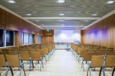 Image of a conference room with symmetrically arranged wooden chairs facing a podium with microphones and a screen.