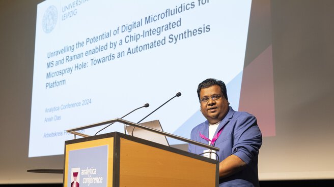 Anish Das from Leipzig University stands on stage behind a lectern at the analytica conference 2024 and gives a presentation. The presentation can be seen in the background.