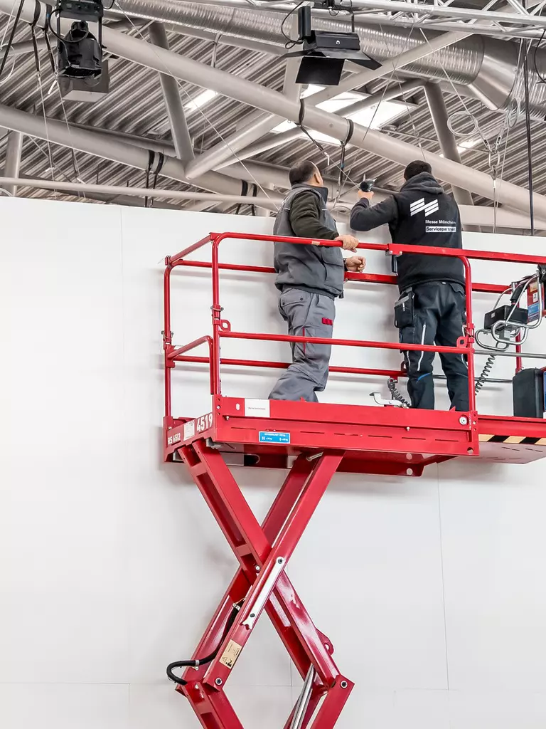 Zwei Servicemitarbeiter der Messe München in Arbeitskleidung stehen auf einer roten Scherenbühne in einer Messehalle.