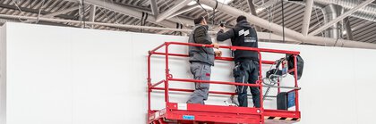 Zwei Servicemitarbeiter der Messe München in Arbeitskleidung stehen auf einer roten Scherenbühne in einer Messehalle.
