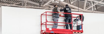 Two Messe München service employees in work clothes stand on a red scissor lift in an exhibition hall.