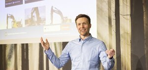 Ein Mann mit kariertem Hemd und Mikrofon hält gestikulierend einen Vortrag vor einer Leinwand mit Bildern von Baumaschinen.