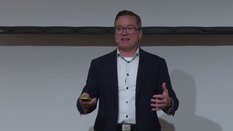 Ein Mann im Anzug und mit Brille spricht und gestikuliert auf einer Bühne während einer Präsentation.