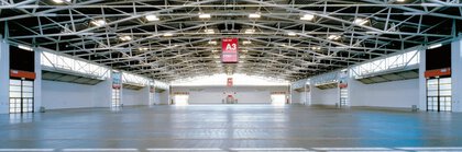 Eine leere Messehalle mit Stahlstreben unter der Decke sowie beleuchteten Fenstern und Türen. In der Mitte hängt ein großes rotes Schild mit der Bezeichnung 