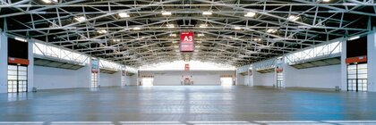 A completely empty exhibition hall with steel struts and windows on the ceiling. In the middle hangs a large red sign with the designation 