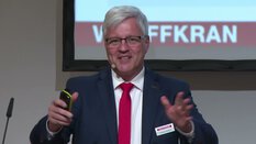 Mann im Anzug und roter Krawatte hält Präsentation, auf dem Bildschirm hinter ihm sind teilweise Texte zu sehen.