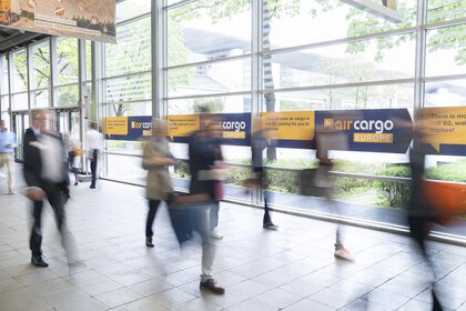 Blurred silhouettes of passing visitors in a connecting corridor with a large window front and signposts to Hall B2 of air cargo Europe.
