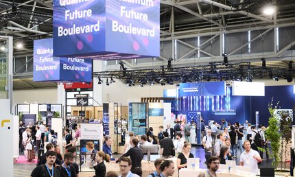 Blick über einen Teilbereich einer Messehalle mit zahlreichen Messeständen, an denen Aussteller und Besucher miteinander ins Gespräch kommen.