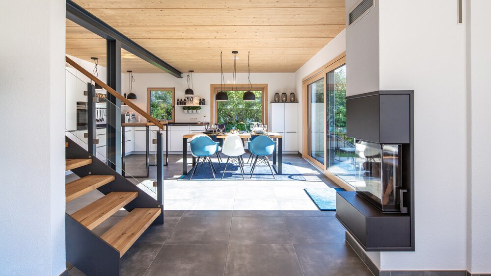 Moderner offener Essbereich mit blauen Stühlen, Holztreppe, großen Fenstern und einem minimalistischen Kamin.