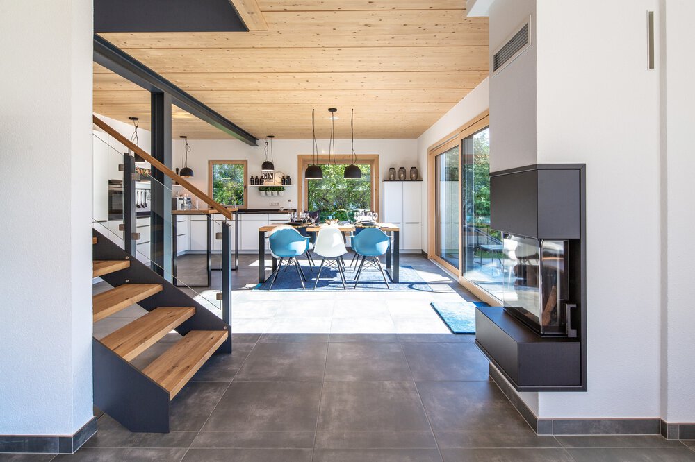 Moderner offener Essbereich mit blauen Stühlen, Holztreppe, großen Fenstern und einem minimalistischen Kamin.