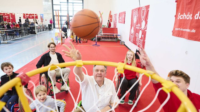 Menschen im Rollstuhl spielen begeistert Rollstuhlbasketball auf einer Messe, ein Ball fliegt Richtung Korb.