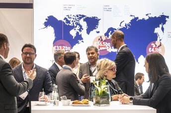 Geschäftsleute im Gespräch auf einer Messe vor einer Weltkarte mit Finanzkennzahlen