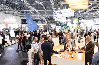 A busy exhibition hall with bustling people and stands, surrounded by banners and posters.