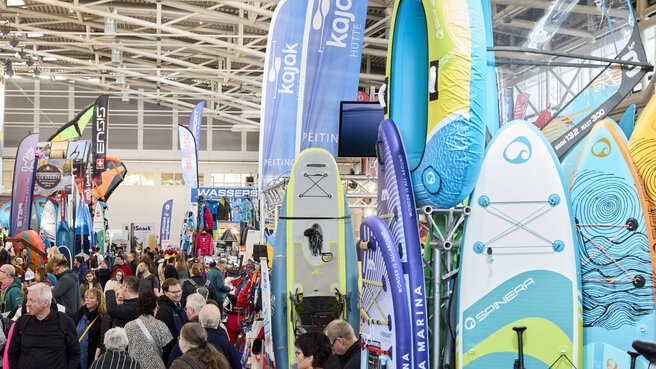 Exhibition stand at the f.re.e trade fair with colorful SUP boards, kayaks and water sports accessories, surrounded by many visitors.