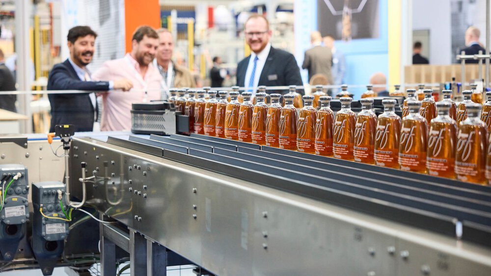 Mehrere Messebesucher betrachten eine große rechteckige Maschine, auf der in Reihen zahlreiche kleine Flaschen mit orangener Flüssigkeit stehen.