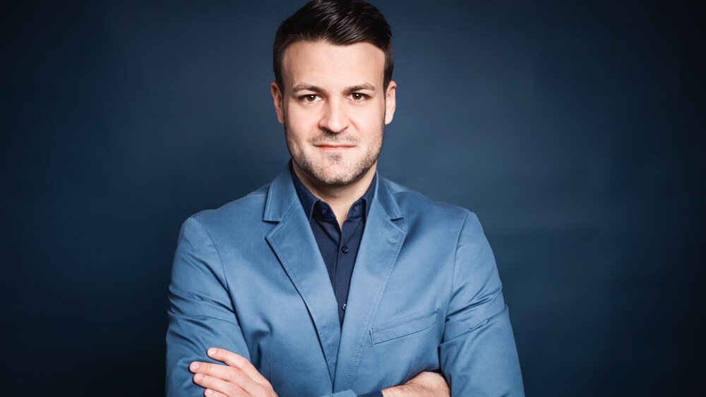 Maximilian Heiler steht selbstbewusst im blauen Anzug mit verschränkten Armen vor einem dunkelblauen Hintergrund.