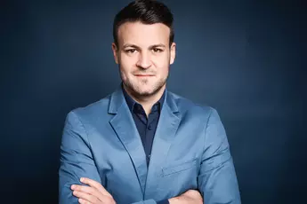 Maximilian Heiler stands confidently in a blue suit with his arms folded in front of a dark blue background.