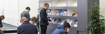 People reading magazines at a public reading stand with signage in German and English.