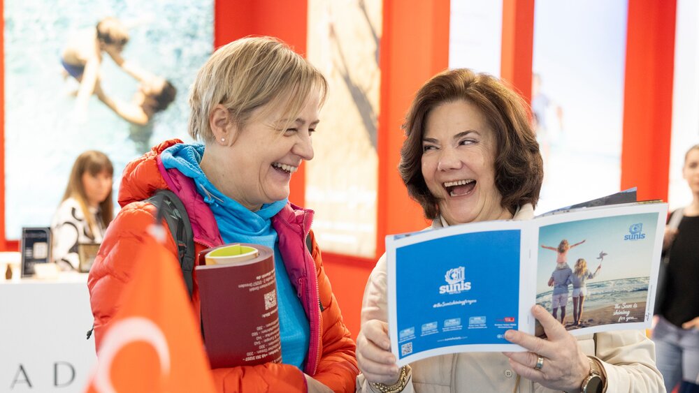 Two smiling women stand together at a stand with a Turkish flag at f.re.e and leaf through a travel magazine.