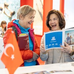 Zwei lachende Frauen stehen gemeinsam an einem Stand mit einer Türkischen Flagge auf der f.re.e und blättern durch ein Reisemagazin.